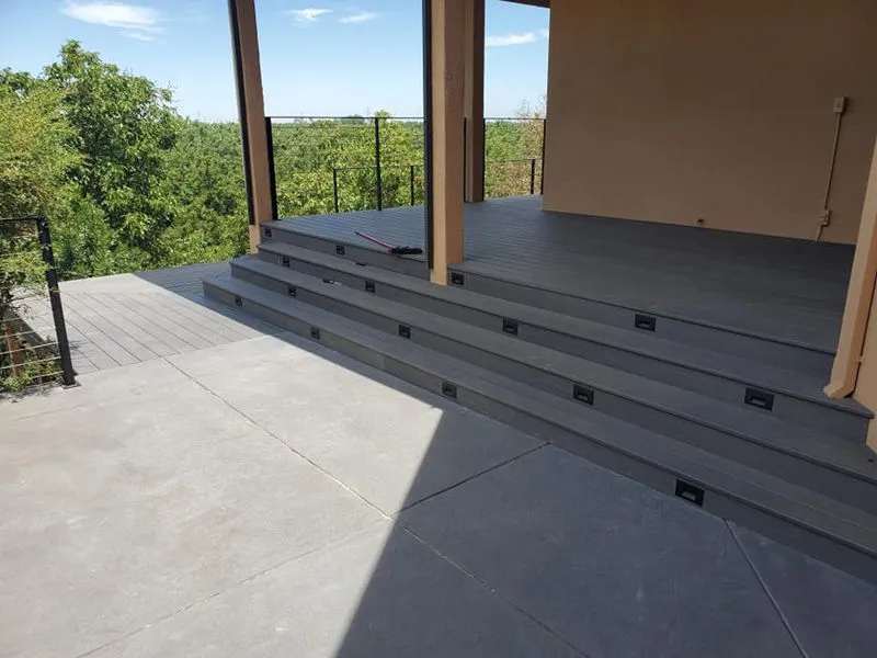 A view of a modern gray deck featuring three wide steps leading up to a covered porch with a wooded background.
