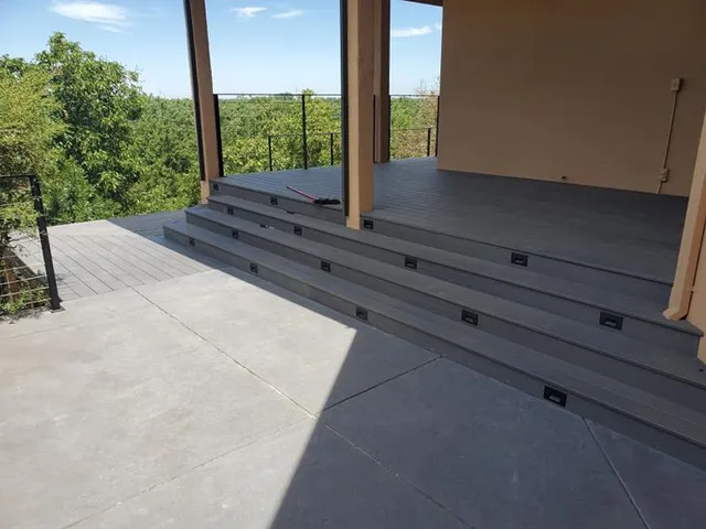 A view of a modern gray deck featuring three wide steps leading up to a covered porch with a wooded background.