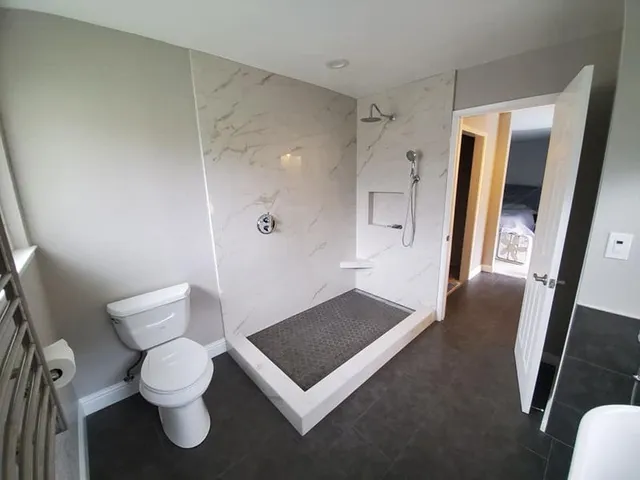 A modern bathroom featuring a walk-in shower with white marble-style walls, a toilet, and an open doorway to a bedroom.
