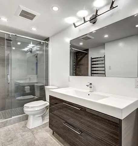 A modern bathroom with a wood-grain vanity, white sink, glass-enclosed shower, toilet, and a large mirror with lighting.