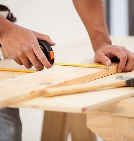 A person uses a yellow tape measure to mark a measurement on a wooden board.