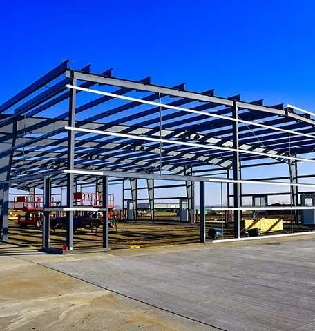 The steel framework of a large, unfinished warehouse stands against a clear blue sky, with construction equipment inside.