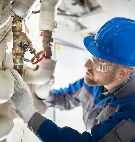 A technician in a blue hard hat, safety glasses, and work gloves adjusts a valve on industrial pipes.