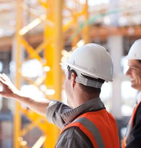 Two construction workers in hard hats and safety vests stand on a job site, with one pointing toward the structure.