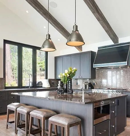 Modern kitchen with dark cabinets, a stone island, four brown bar stools, and two metal pendant lights under wood beams.
