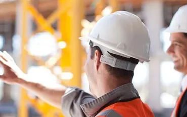 Two general contractors in hard hats and safety vests stand on a construction site, with one pointing forward.
