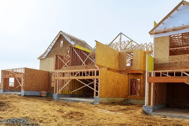A row of unfinished wooden houses under construction at a sandy site with exposed framing and plywood panels.