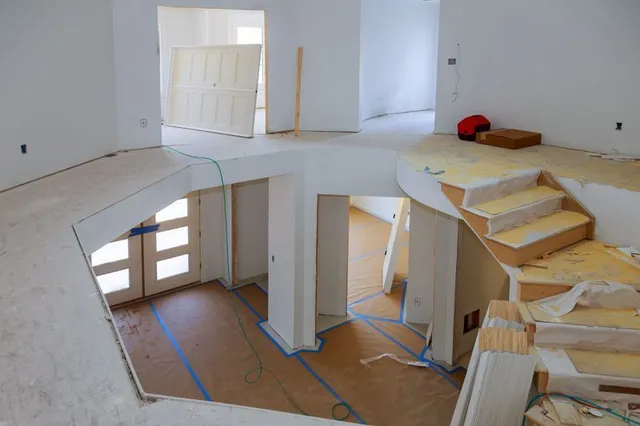 An interior view of a two-story home under construction, showing a staircase, unfinished floors, and white walls.