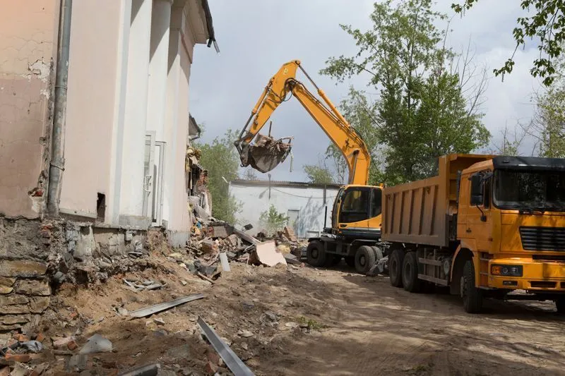 A yellow excavator demolishes a damaged building beside a dump truck on a dirt-covered construction site.