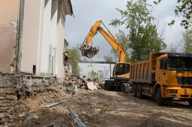 A yellow excavator demolishes a damaged building beside a dump truck on a dirt-covered construction site.