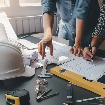 Engineers work at a table with blueprints, a hard hat, measuring tools, and a laptop to review technical drawings.