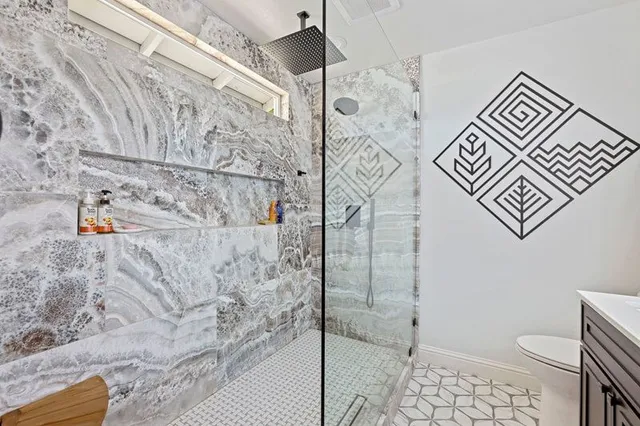 A modern bathroom with a glass-enclosed shower, marbled stone walls, a geometric wall decal, and patterned floor tiles.