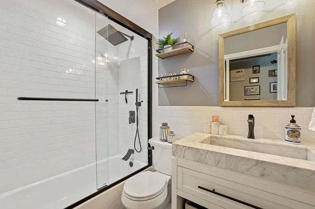 Modern bathroom with a white tiled shower, glass door, vanity with a stone countertop, toilet, and wooden-framed mirror.