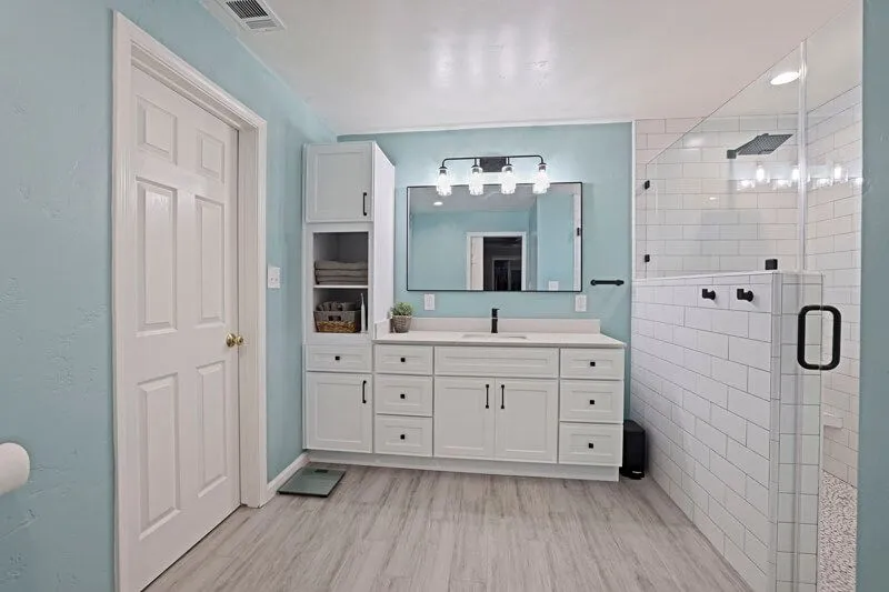 Bathroom with light blue walls, white cabinetry, a single vanity mirror, and a white tiled glass-enclosed shower.