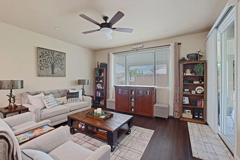 A living room with a beige sofa, two armchairs, a wooden coffee table, two bookcases, and a ceiling fan.
