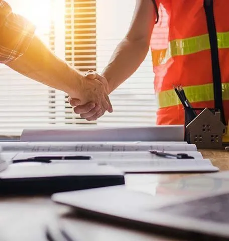 Two people shaking hands over blueprints at a desk with a small model house in an office setting.