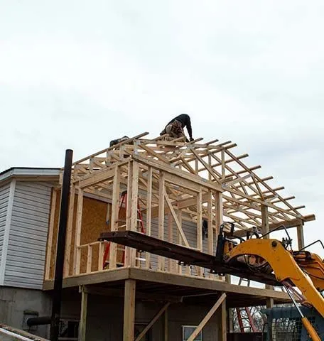 A worker climbs on the wooden roof trusses of a house extension frame under construction, with a yellow excavator nearby.