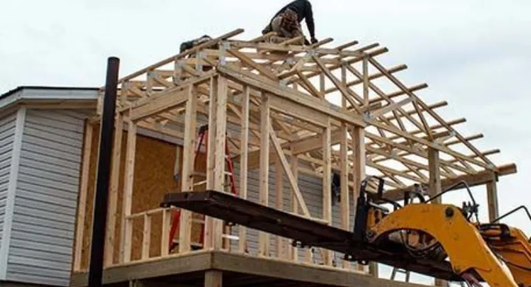 A construction worker builds a wood-framed roof structure on an addition attached to the side of a house.
