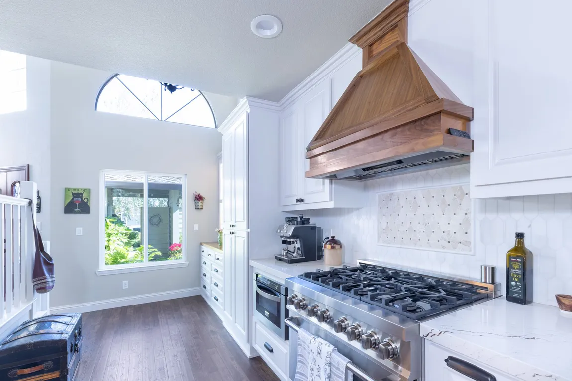 A kitchen featuring white cabinets, a prominent wooden range hood over a gas stove, white counters, and dark wood floors.