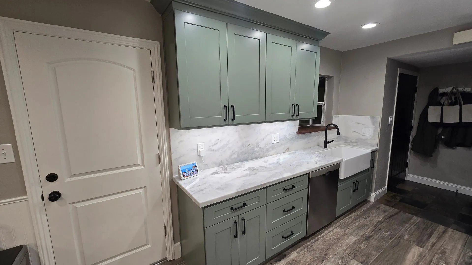 A kitchen area with sage green cabinets, white marble countertops, a white farmhouse sink, and gray plank flooring.