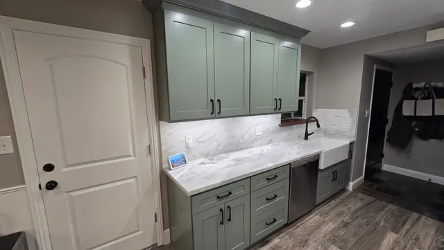 A kitchen area with sage green cabinets, white marble countertops, a white farmhouse sink, and gray plank flooring.