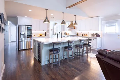 A bright kitchen with white cabinets, stainless steel appliances, a center island with four stools, and dark wood floors.
