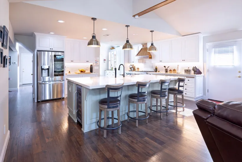 A bright kitchen with white cabinets, stainless steel appliances, a center island with four stools, and dark wood floors.