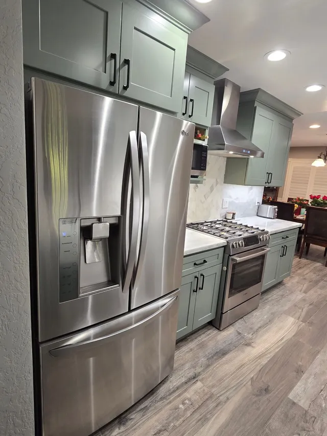 A modern kitchen features sage green cabinets, stainless steel appliances, a range, and light wood-grain flooring.