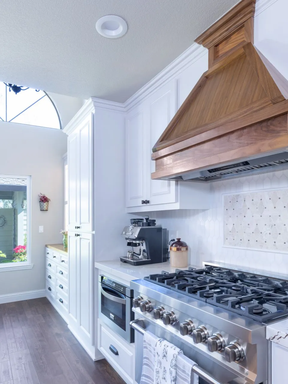 A modern kitchen featuring white cabinets, a prominent wooden vent hood, a stainless steel gas range, and wood flooring.