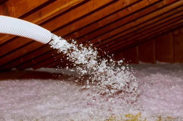 A hose blows loose-fill insulation into a wooden attic space.