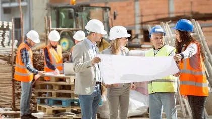 A group of people wearing safety helmets and vests stand at a construction site, reviewing a large set of blueprints.