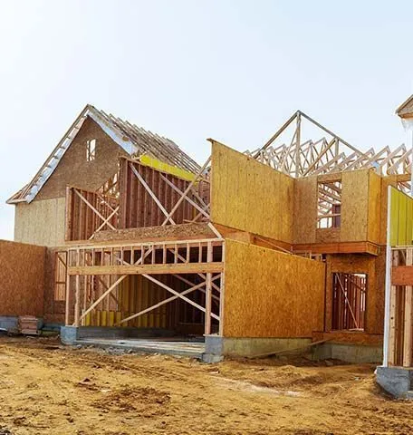 A wood-framed house under construction featuring exposed studs, plywood sheathing, and roof trusses against a pale sky.