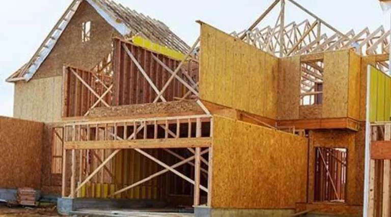 A house under construction showing the wooden frame, roof trusses, and plywood wall sheathing.