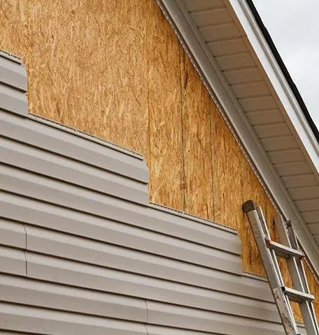 Tan vinyl siding is being installed over an exposed plywood exterior wall on a house, with a ladder leaning against it.