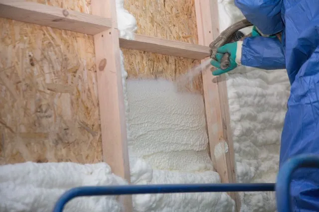 A person in protective gear sprays expanding foam insulation onto the wooden frame of a wall.
