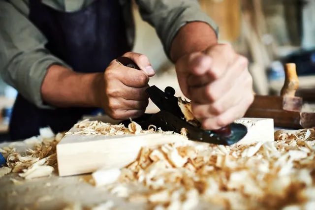 A carpenter uses a handheld plane to smooth a piece of light wood, surrounded by wood shavings in a workshop.